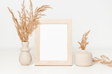 Empty wooden photo frame with beige ceramic vase set and pampas grass on white wooden table. Mockup...
