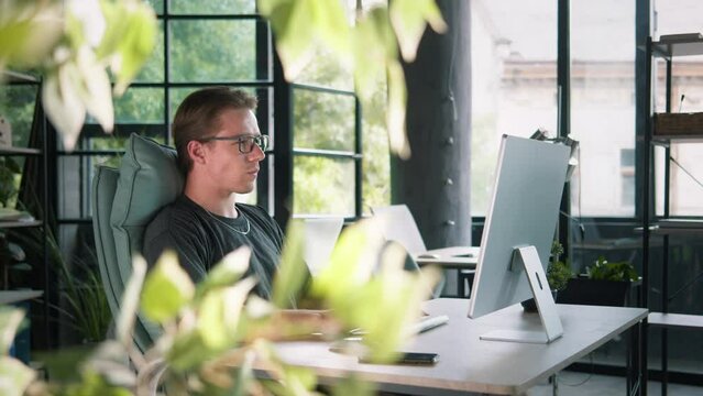 Young caucasian male working at the desktop computer in a comfy informal workplace. Thinking about a task, having an idea, typing on a keyboard