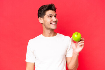 Young caucasian man with an apple isolated on red background looking up while smiling