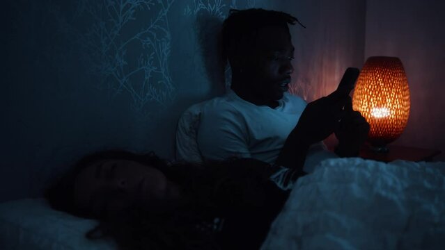 Man smiles and looks over at partner while they lay in bed at night using a smart phone