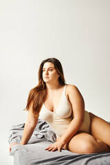 brunette plus size woman with natural makeup and long hair wearing beige bodysuit and posing on bed...