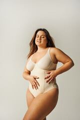 brunette and curvy woman with tattoo wearing beige bodysuit and standing with hands on waist on grey background, body positive, figure type, body positivity movement, closed eyes
