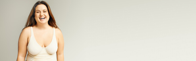 Obraz premium portrait of happy and curvy woman with plus size body posing in beige bodysuit while laughing on grey background, body positive, figure type, looking at camera while standing in studio, banner