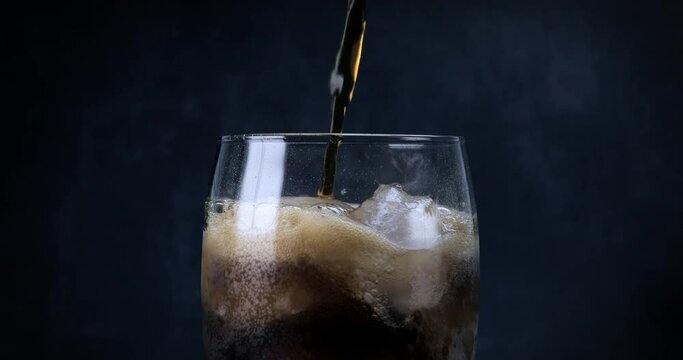 A cool glass of cola drink with ice, bubbles and fizz. Fresh cold sweet drink poured over ice cubes. Cola glass with summer refreshment on black. Slow motion