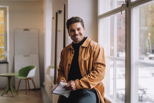 Portrait of smiling male PhD student holding diary sitting by window in innovation lab