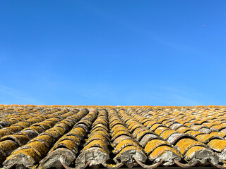 Old, rusty and battered tiled roof with yellow moss on top. Blue sky. Roof with brick roof of an...
