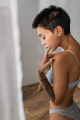 Fototapeta na wymiar side view of young captivating woman with short brunette hair and tattooed arm touching face while standing in lace panties and bra near white curtain on blurred foreground in bedroom