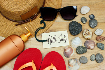summer concept, sun cream and slipper with hello July text on tag and stones. seashell hat on wood table
