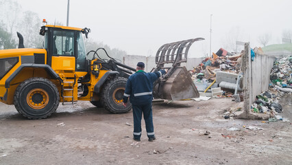 Yellow skid steer loader moving wooden waste material, shaking out a scrap grapple on the garbage heap in the materials recovery facility