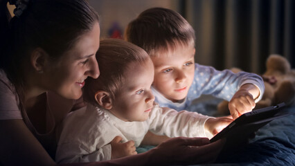 Fototapeta premium Portrait of baby boy using tablet computer with brother and mother in bed at night. Family having time together, parenting, happy childhood and entertainment.