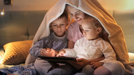 Happy family sitting under blanket and using tablet computer at night. Family having time together, parenting, happy childhood and entertainment.