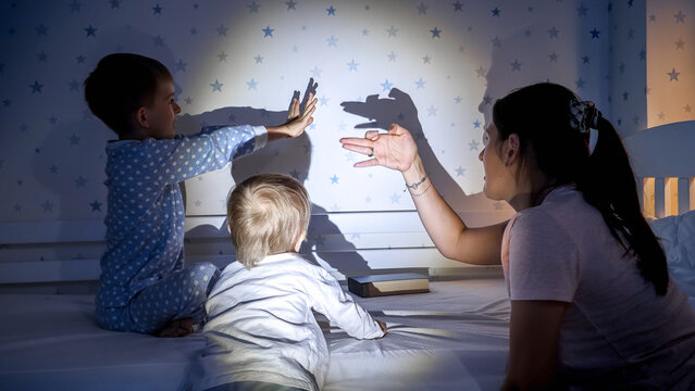 Cheerful kids with mother playing with shadows from the flashlight on wall at night. Family having time together, parenting, happy childhood and entertainment.