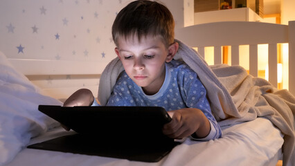 Dolly shot of little boy in pajamas playing on tablet computer before going to sleep. Children education, development, kids using gadgets secrecy, privacy