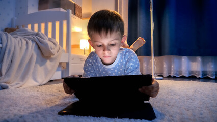 Cute boy in pajamas lying on floor in his room and using tablet computer before going to bed. Children education, development, kids using gadgets secrecy, privacy