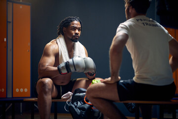 Powerful boxer in sweats resting on bench after workout, talking with his coach, getting...