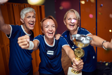 Lovely team of senior and young women celebration win with golden cup together in locker room.