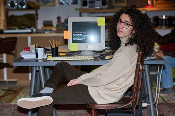 Portrait of young programmer with curly hair looking at camera sitting at his workplace with...
