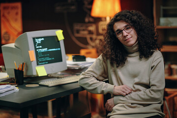 Portrait of young developer with long curly hair looking at camera while sitting at his workplace...