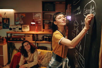 Young girl drawing business plan on blackboard while working with her team over new computer...