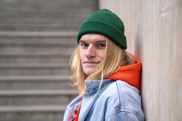 Urban portrait of happy millennial hipster guy with long blond hair smiling at camera, leaning on...
