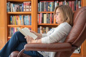 Senior white woman sitting comfortably in a relaxing chair while reading a book.