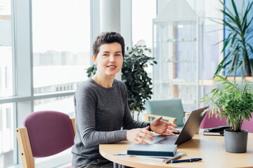 Neutral gender middle aged woman with no make-up in casual clothing using laptop while working indoors on workstation in an open space office, coworking, library. Remote workplace, selective focus.