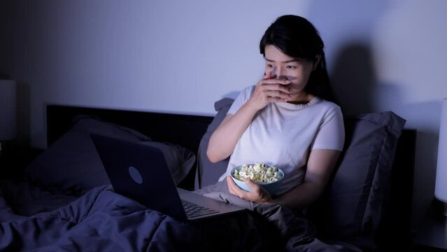 Asian woman eating popcorn while watching a scary movie on her laptop while sitting in bed.