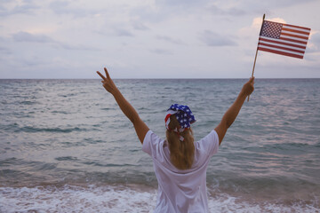 A blonde girl is standing on the ocean. In his right hand he holds an American flag, and his left hand shows 2 fingers up. Soft focus