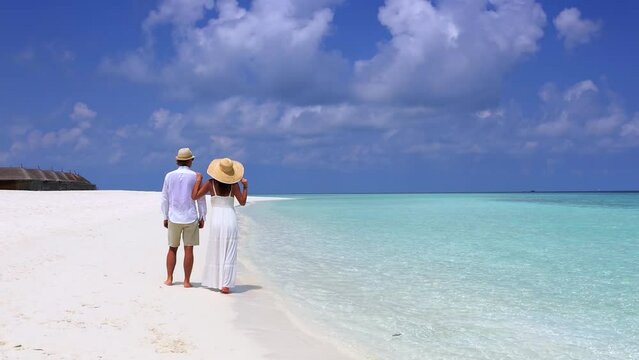 Happy couple in white clothing and with hats stands a tropical beach with turquoise sea in the Maldives islands and enjoys the view