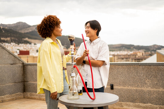 A couple of multiethnic friends are having a few beers and smoking a hookah. The African girl with the afro hair and the Korean boy talk happily. Concept of multiracial couple.