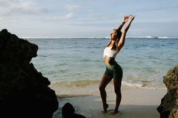Slender attractive young woman during a meditation dance, raising her hands above her head in a cave among the rocks on the beach near the ocean