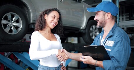Happy automotive mechanic man in uniform shaking hands with women client at auto repair shop. Car...