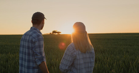 A young couple of farmers watch how a tractor works in the field. Stand side by side against the...
