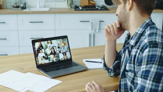 Side view of caucasian guy sitting in kitchen at home talking by video conference with group of multiracial successful colleagues discuss company promotion, advertising, financial strategy,new project