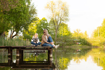 Caucasian Mother And Son Sit On Wooden Dock Enjoying Lake View In Summer Time. Boy Throws Rocks Into Water. Mom's Support And Care. Motherhood, Family Leisure Time. Children's Day. Horizontal Plane