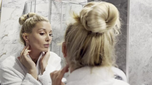 mid age adult blonde woman wears bathrobe and towel in bathroom applying rejuvenating tightening  antiage face skin care cream treatment.  Daily beauty routine.
