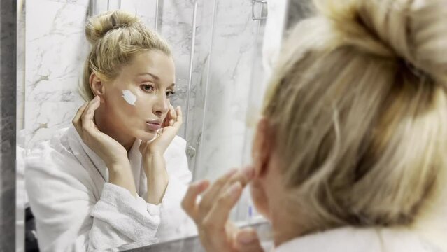 middle aged woman touching face skin looking in mirror reflection.  Smiling mature woman pampering, healthy moisturized skin care, aging beauty,  skincare treatment cosmetics concept.