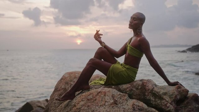 Androgynous person of color stands in boutique open dress on rock above ocean coastline on sunset. Trans sexual ethnic fashion model in skimpy outfit wears golden jewellery, posing like Oshun goddess.