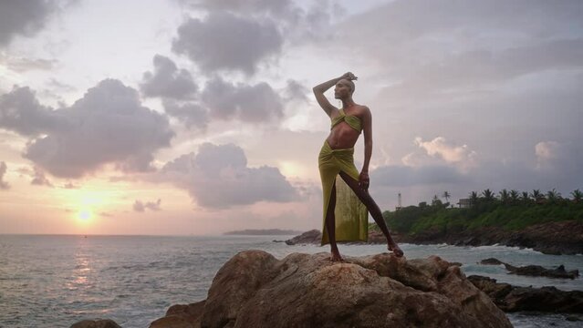 Divine non-binary black person in botique dress, brass jewelry poses on rocky hill top above dramatic ocean sunset skyline. Queer lgbtq fashion model in open outfit fluttering in air. Pantheon concept