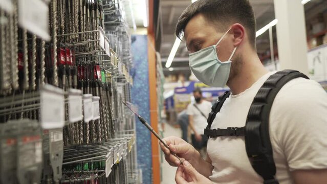 Caucasian man in medical mask selects goods drill bit for construction tool in hardware store. Male choice metal drill set for drilling machine in building supermarket during the covid 19 pandemic.