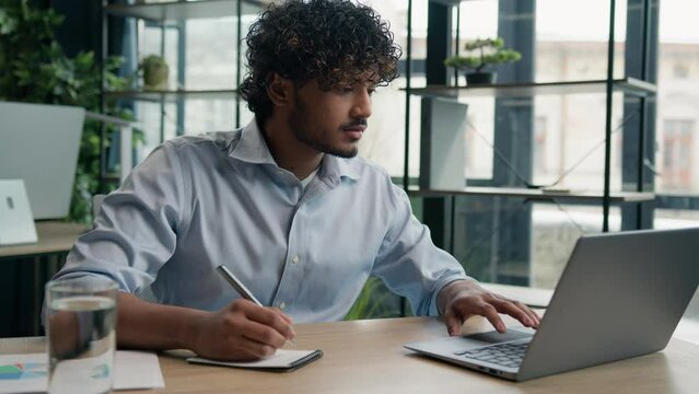 Indian Arabian man accountant writer business entrepreneur businessman write notes in notebook latino guy writing ideas report paperwork on office table studying online with laptop educational course