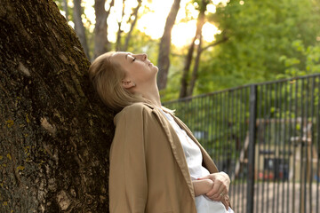 Tired White Woman Leans To Tree, Closed Her Eyes In Summer Time on Sunset. Rest And Recharge in Nature, Self Awareness, Leisure Time, Mental Health. Copy Space For Text Horizontal Plane