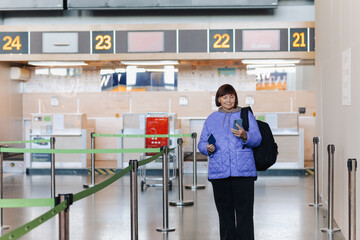 Fototapeta na wymiar Middle aged woman traveler with backpack holds passport, uses cellphone at public airport terminal after check in at airline services desks. Active retirees. Travelling, vacation, tourism concept.
