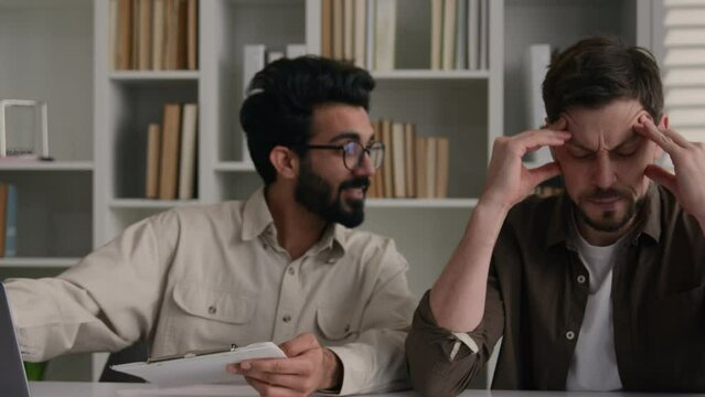 Overworked tired sick man suffer headache ignoring obsessive annoying colleague disturb talk smile discuss project with laptop two multiethnic businessmen diverse men solve teamwork problem in office