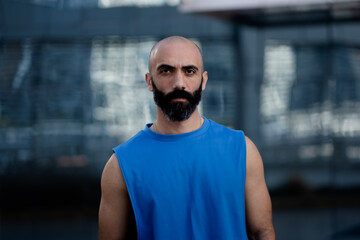 Looking directly at the camera, a man poses for a portrait outdoors. He is wearing a sleeveless shirt that shows off his lean physique. He has strong Middle Eastern features.