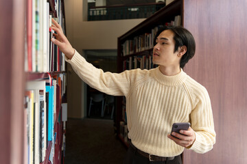 Portrait of asian man choosing book in university library, holding mobile phone, modern education