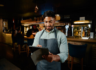 Young biracial man in apron using digital tablet while working as waiter in bar
