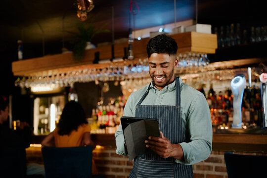 Happy young biracial man in apron using digital tablet while working as waiter in restaurant