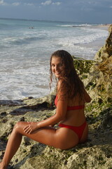 Fototapeta na wymiar A woman with curly hair in a red swimsuit sits on a rocky beach.