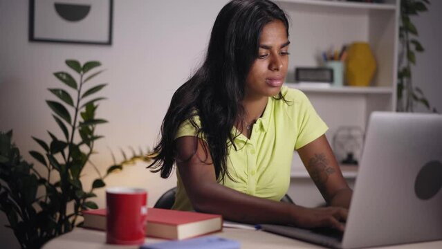Young Indian woman using laptop in home office. Serious girl typing on computer and taking notes. Concentrated people studying for final exam. Concept of person preparing competitive examination. 